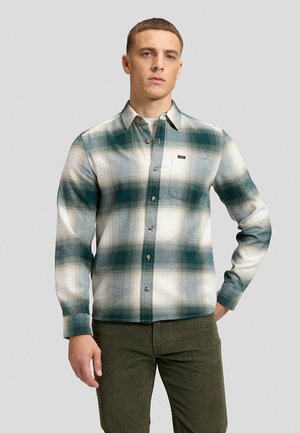 Uomo con camicia a quadri verde e bianca con taschino sul petto e pantaloni verde oliva, in piedi su uno sfondo semplice.