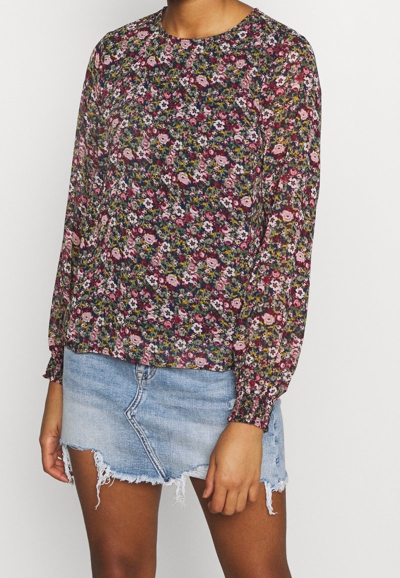 Blommig långärmad blus i mörka färger med rund halsringning, smockade manschetter och ett lätt, genomskinligt tyg över en denim minikjol.