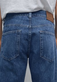 Jean en denim bleu à texture lisse, avec deux poches arrière et une étiquette en cuir à la taille. Design classique à jambes droites.