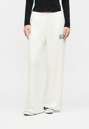 Person iført løstsiddende hvide bukser med "GAP"-logo på venstre lår, sort langærmet top og sort-hvide sneakers.