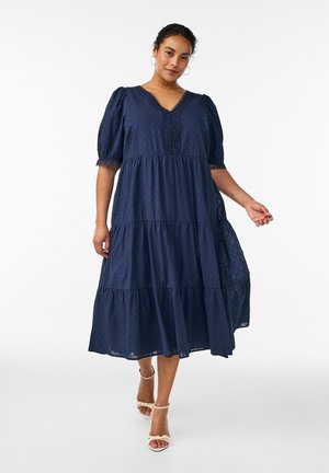 Kvinde iført en marineblå midi-kjole med lag, puffede ærmer og blonderkant, kombineret med hvide ankelrem-sandaler, stående på en hvid baggrund.