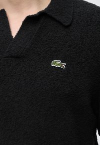 Μαύρο πόλο με υφή και πράσινο κεντημένο λογότυπο κροκόδειλου στην αριστερή περιοχή του στήθους, που φοριέται από ένα άτομο.