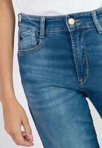 Jeans de mezclilla azul con un ajuste slim, que presentan un cierre de botón dorado, cinco bolsillos y costuras en contraste. Textura suave y cintura media.