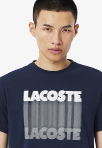 Camiseta de algodón azul marino con un gráfico en blanco audaz de "LACOSTE" con detalles de líneas verticales. Manga corta y cuello redondo.