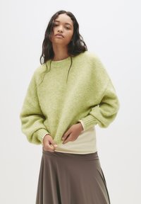 Mlada žena prilagođava svijetlozelenu oversized majicu, nosi bež top i lepršavu smeđu suknju uz jednostavnu bijelu pozadinu.