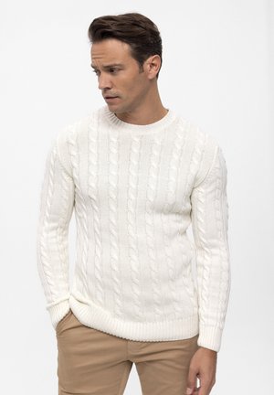 Homme portant un pull en maille torsadée blanc et un pantalon marron clair, regardant vers le bas et sur le côté, les mains dans les poches, sur un fond uni.