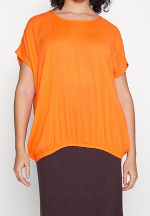 Femme portant un haut orange ample à manches courtes associé à une jupe côtelée foncée, vue du cou aux hanches.