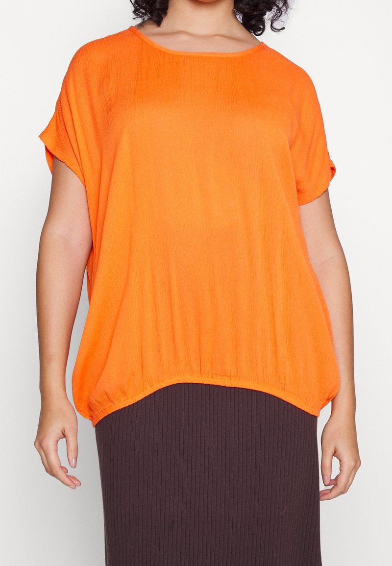 Femme portant un haut orange ample à manches courtes associé à une jupe côtelée foncée, vue du cou aux hanches.