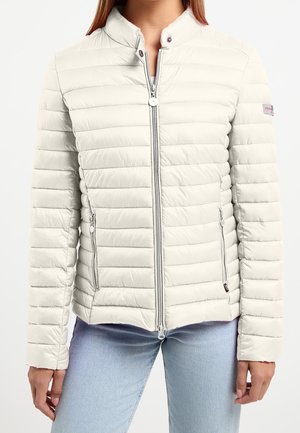 Femme portant une veste matelassée beige clair avec une fermeture éclair frontale et des poches zippées, associée à un jean bleu clair.