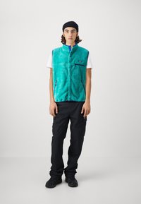Turkis fleece vest med et teksturert mønster, glidelåslommer, blå kant, brukt over en hvit t-skjorte, sammen med svarte cargo bukser og sko.