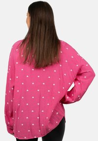 Donna con lunghi capelli marroni, che indossa una camicia rosa larga con motivi a cuori bianchi, con una mano sulla vita, rivolta di spalle rispetto alla macchina fotografica.