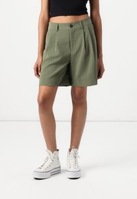 Shorts plissés en kaki fabriqués en tissu léger, avec une fermeture à bouton sur le devant, des poches latérales et une coupe décontractée. Portés avec des baskets blanches.