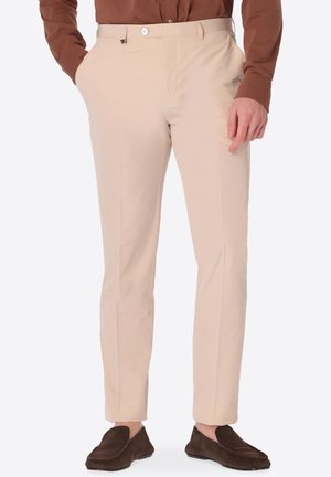 Uomo che indossa pantaloni beige slim fit con le mani nelle tasche, abbinati a mocassini marrone scuro e una camicia marrone a maniche lunghe.
