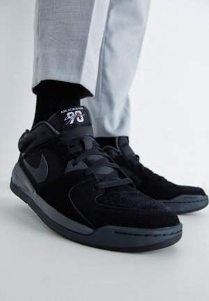 Persona con zapatillas Air Jordan negras con suelas grises y pantalones grises, de pie sobre un suelo blanco con un fondo blanco.