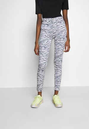 Mujer vestida con pantalones skinny de cintura alta en estampado de cebra gris y blanco, con una blusa negra y zapatillas de deporte en neón amarillo, de pie contra un fondo blanco.