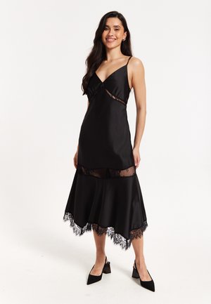 Black satin dress with spaghetti straps, featuring a sheer lace trim detail and flared hem with lace edging. Smooth texture and fitted silhouette.