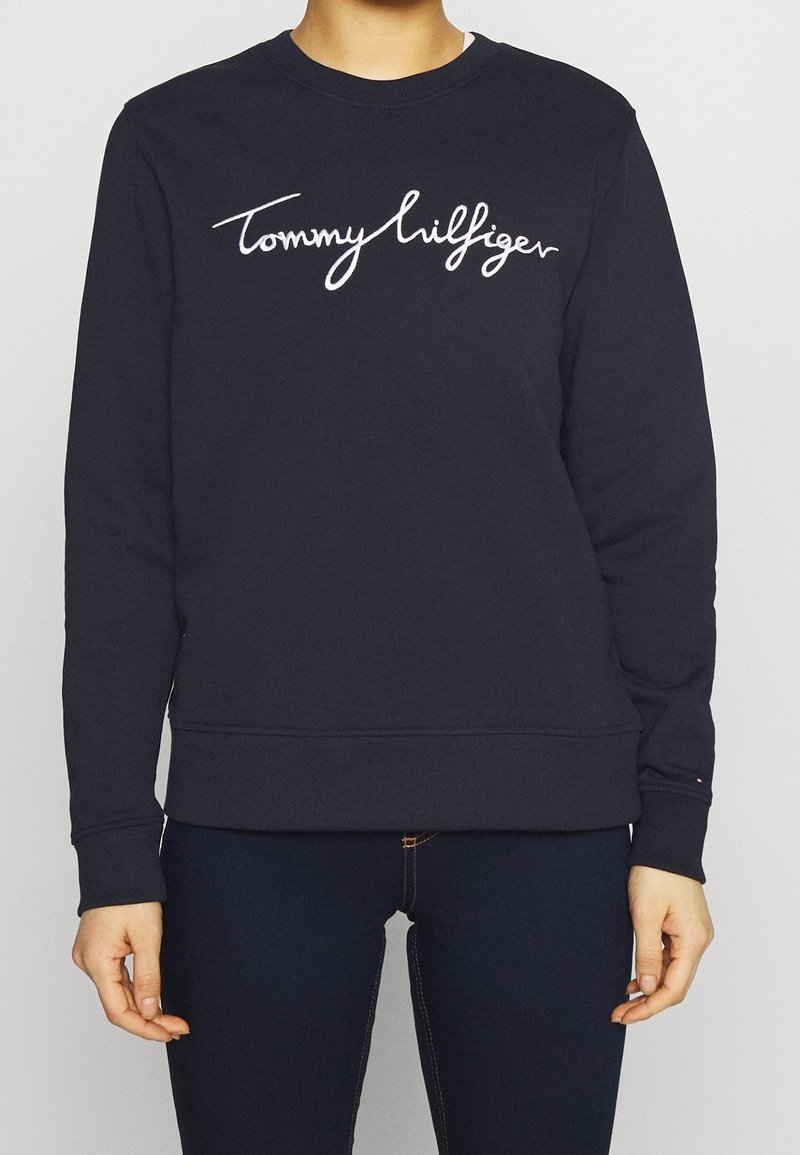 Personne portant un sweat-shirt Tommy Hilfiger bleu marine avec un logo en écriture blanche sur la poitrine et un jean bleu foncé.