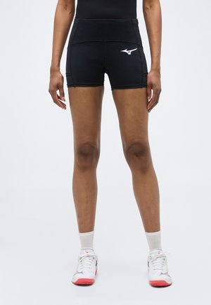 Άτομο που φοράει μαύρο αθλητικό σορτς με λογότυπο, λευκές κάλτσες και λευκά αθλητικά παπούτσια με ροζ σόλες, στέκεται σε απλό ανοιχτόχρωμο φόντο.