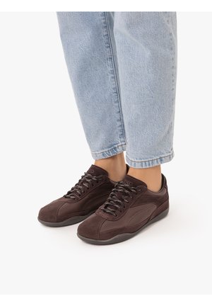 Osoba w jasnoniebieskich dżinsach i ciemnobrązowych sznurowanych sneakersach stojąca na białej powierzchni.