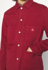 Veste en coton rouge avec un devant à bouton, deux poches poitrine et deux poches latérales. Présente un petit patch logo et une finition solide et texturée.