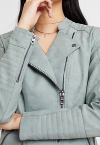 Veste de motard en similicuir gris clair avec fermeture éclair sur le devant, détails matelassés aux épaules et aux coudes, et col montant.