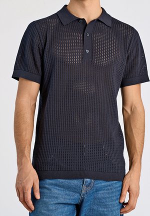 Navy blue knitted polo shirt featuring a textured, open pattern, ribbed collar and cuffs, and three-button placket. Paired with casual jeans.