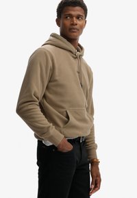Cafékleurige fleece hoodie met een kangoeroezak en een capuchon met trekkoord, voorzien van subtiele branding. Gecombineerd met donkere jeans en een riem.