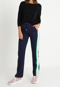 Marina joggers med grön sidostrimma och vit logotyp, elastisk midja med dragsko, ihopparat med en svart topp och rosa sneakers.