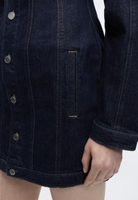 Σκούρο μπλε denim μπουφάν με μεταλλικά κουμπιά και κάθετη τσέπη, εμφανίζεται από τη μέση έως το ύψος του χεριού σε άτομο με ανοιχτόχρωμο δέρμα.