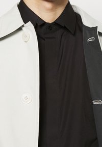 Cappotto grigio chiaro con colletto a punta, grandi bottoni bianchi, indossato sopra una camicia nera. L'interno mostra dettagli di cucitura e chiusura con bottoni.