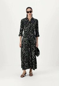 Juoda suknele su spaudiniu, su mygtukų užsegimu priekyje, trijų ketvirčių rankovėmis ir laisva siluetu. Nešiojama su juodais šleptais sandalais ir rankine.