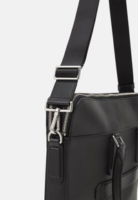 Sort læder skuldertaske med en struktureret overflade, sølvfarvet hardware, lynlåslukning og justerbar strop. Har et slankt, rektangulært design.