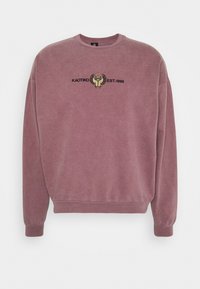 Plommonfärgad sweatshirt med ribbad mudd och nederkant. Har broderad design med texten "KAOTIKO EST.1999" och ett grafiskt accent. Mjuk textur.