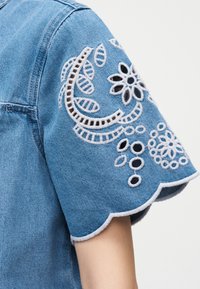 Chemise à manches courtes en denim bleu avec des motifs floraux ajourés brodés en blanc et un bord festonné sur le bras d'une personne.