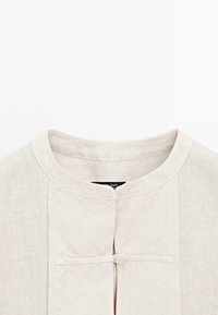 Jasnobeżowa lniana bluzka z okrągłym dekoltem, zapięciem na guziki z przodu oraz teksturowaną tkaniną. Prosty, minimalistyczny design.