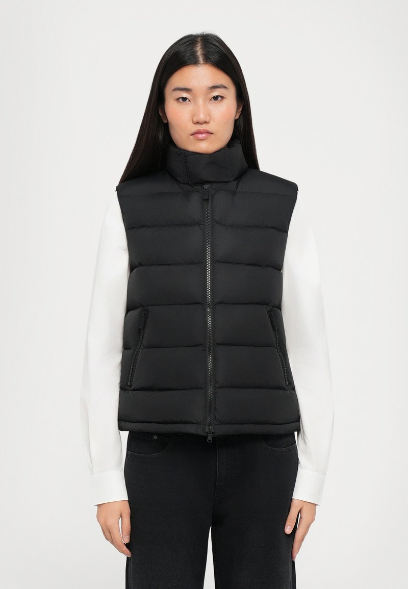 Giovane donna con lunghi capelli scuri che indossa un gilet nero imbottito sopra una maglietta bianca a maniche lunghe e pantaloni neri, in piedi su uno sfondo neutro.