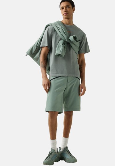 Mladý muž, který má na sobě tlumené zelené šortky, odpovídající oversized tričko, tenisky a přes jedno rameno uvázanou bundu, stojící na světlém pozadí.