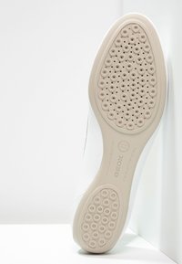 Vit sneakersula med cirkulära ventilationshål och texturerad yta. Har en slät kant och präglad logotyp.