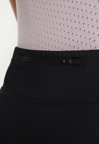 Schwarze Sportleggings mit glatter Textur und breitem Bund, ausgestattet mit einer kleinen Reißverschlusstasche und einem helllila Top mit Lochmuster.