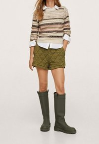 Stivali di gomma verde oliva, pantaloni corti imbottiti verde oliva e un maglione di maglia a righe in beige, marrone e rosa, layered su una camicia bianca.
