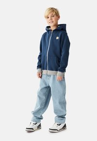 Marineblå lynlåshættejakke med grå accenter, med en forlomme, båret med lyseblå løstsiddende jeans og hvide sneakers med sorte detaljer.