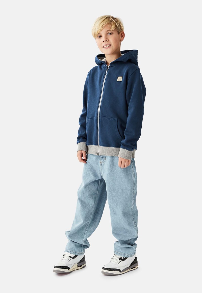 Marineblå lynlåshættejakke med grå accenter, med en forlomme, båret med lyseblå løstsiddende jeans og hvide sneakers med sorte detaljer.