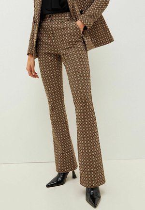 Frau trägt braune Anzughose mit geometrischem Muster und passenden Blazer, kombiniert mit schwarzen spitzen Stiefeln mit Absatz, steht vor einer weißen Wand.