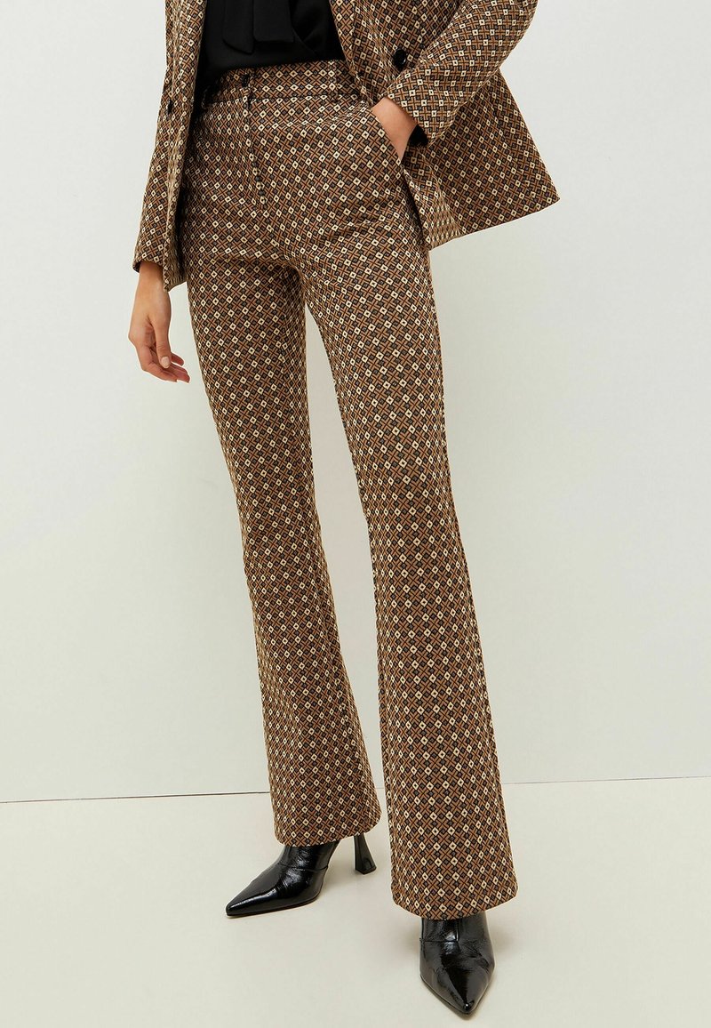 Femme portant un pantalon de costume à imprimé géométrique marron et un blazer assorti, associés à des bottines noires à talons et bouts pointus, se tenant contre un mur blanc.