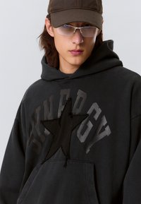 Schwarzer Hoodie mit einem Stern-Patch, mit "SYNERGY" in dunkler Schrift. Weicher Stoff mit einer Kängurutasche und Kapuze mit Kordelzug.