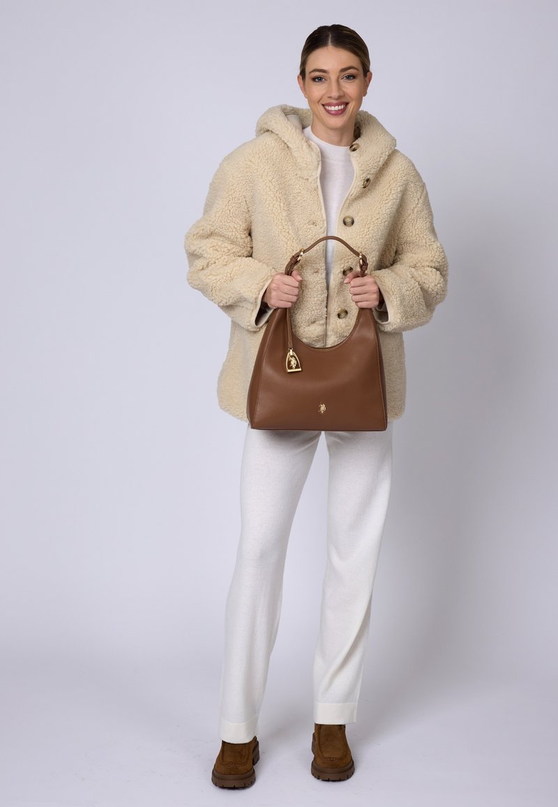 Borsa a tracolla in pelle marrone con silhouette arrotondata e dettagli in oro. Cappotto teddy beige testurizzato con cappuccio e grandi bottoni.