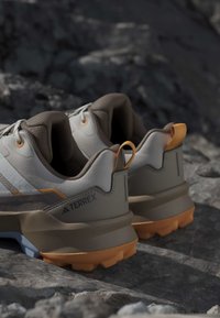 Paire de chaussures de trail running Adidas Terrex beige et orange placées sur un terrain rocheux, mettant en avant une semelle robuste et des languettes au talon.