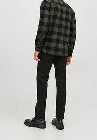 Grün-schwarz kariertes Flanellhemd, zugeknöpft, kombiniert mit schwarzer Hose und schwarzen Knöchelstiefeln, von hinten gesehen. Glatte Stoffstruktur.