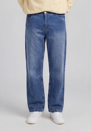 Personne portant un jean bleu ample et des baskets blanches sur un fond clair uni, visible de la taille aux pieds.