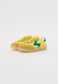 Yellow suede sneakers with green accents, featuring a thick translucent sole and textured laces. Bolt-shaped detail on the sides.
